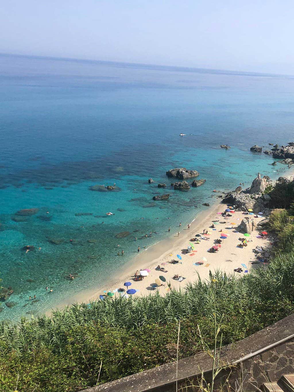 A small beach with clear blue water and beach umbrellas, surrounded with green vegetation. 