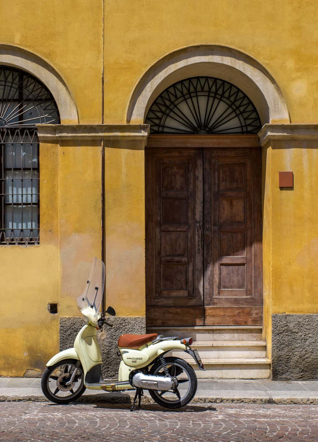 A vintage moped scooter parked on. cobbled street in front of a yellow building with big wooden doors in Italy. 