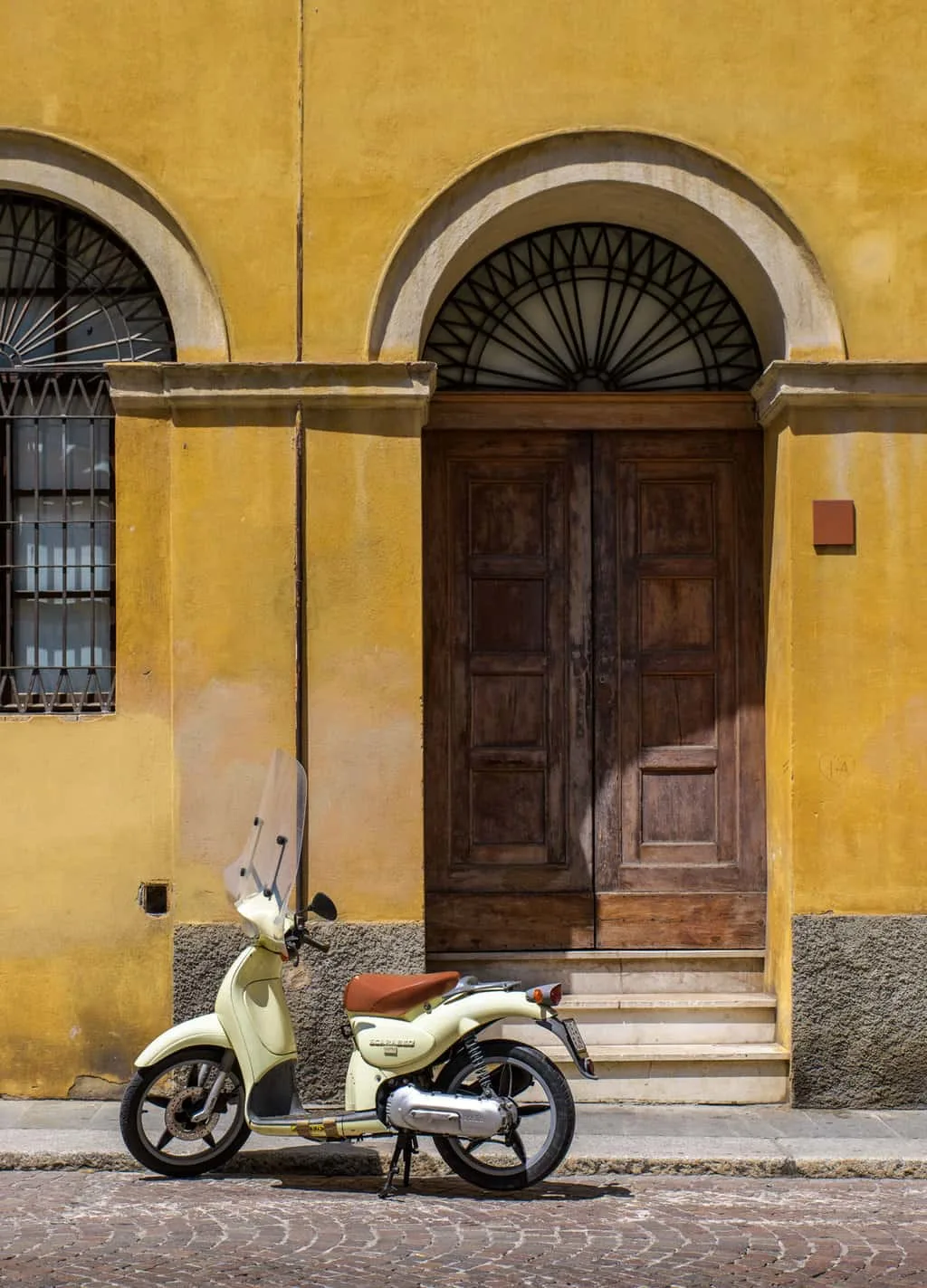 A vintage moped scooter parked on. cobbled street in front of a yellow building with big wooden doors in Italy.