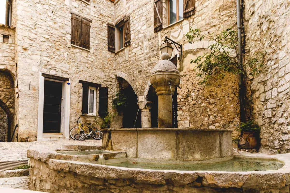 A medieval stone fountain in a small town square surrounded by stone buildings in Peille France.