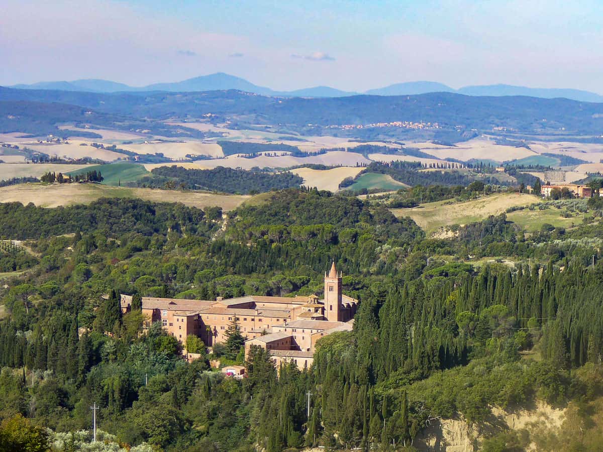 Rolling hills of Tuscany with a large red brick monastery in the centre surrounded by trees. 