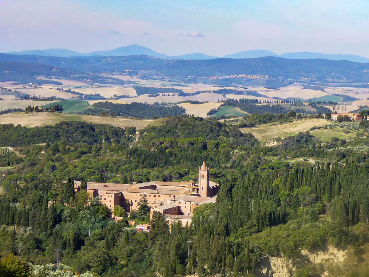 Rolling hills of Tuscany with a large red brick monastery in the centre surrounded by trees.