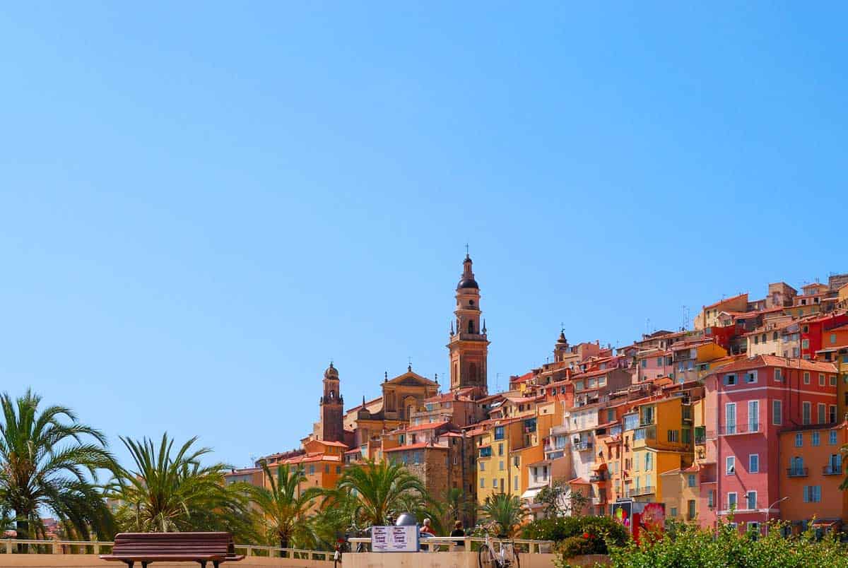 Brightly coloured houses cascade down the hill punctuated with church spires against a blue sky and palm trees in the foreground in the village of Ventimiglia. 