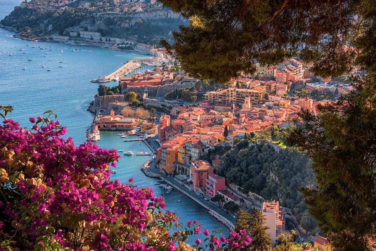 Looking over the old town and harbour of Villefranche-Sur-Mer with blue water and orange hued buildings. Pink Bougainvillea is in the foreground. 