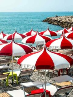 Red and white umbrellas and sun lounges line the pebble beach in Cagnes-sur-Mer overlooking turquoise blue water on a sunny day.