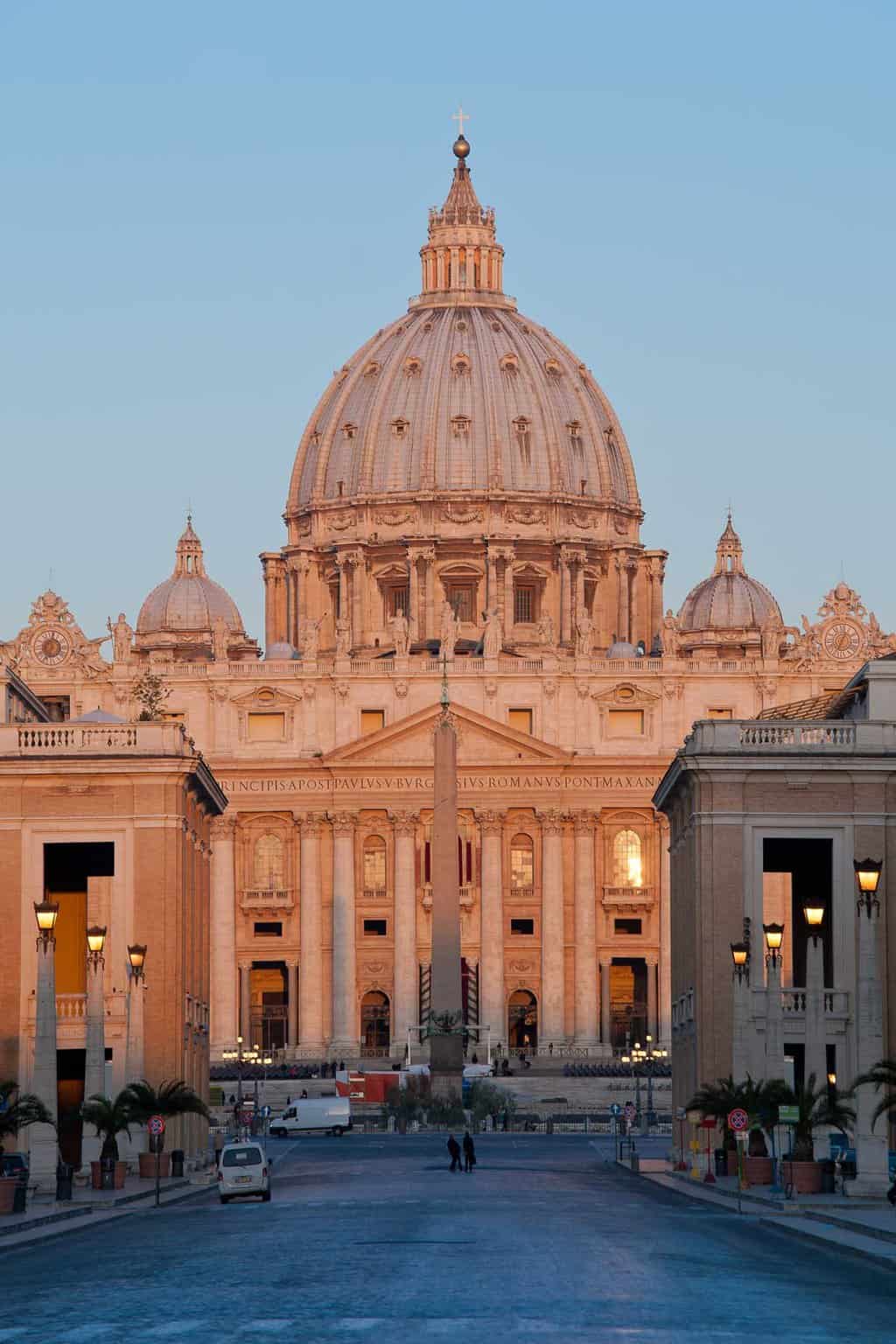 The exterior of St Peter's Basilica bathed in a light pink glow at sunrise.