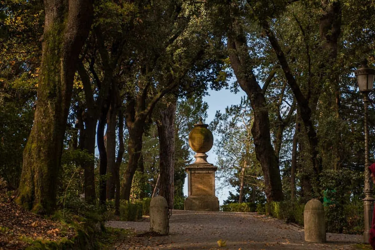 A large decorative urn on a stand at the end of a path lined with shady trees in the Vatican Gardens.