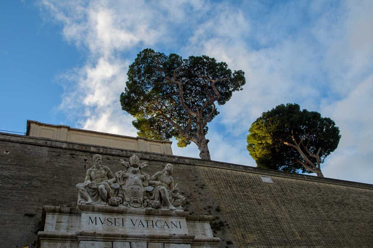 Statues above the Vatican Museum sign written in Latin on stone on a tall wall with trees rising above the wall.