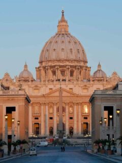 The exterior of St Peter's Basilica bathed in a light pink glow at sunrise.