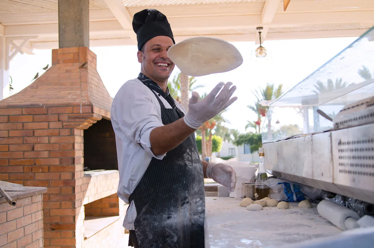 A smiling chef in a chefs hat tossing pizza dough in front of an outdoor pizza oven.