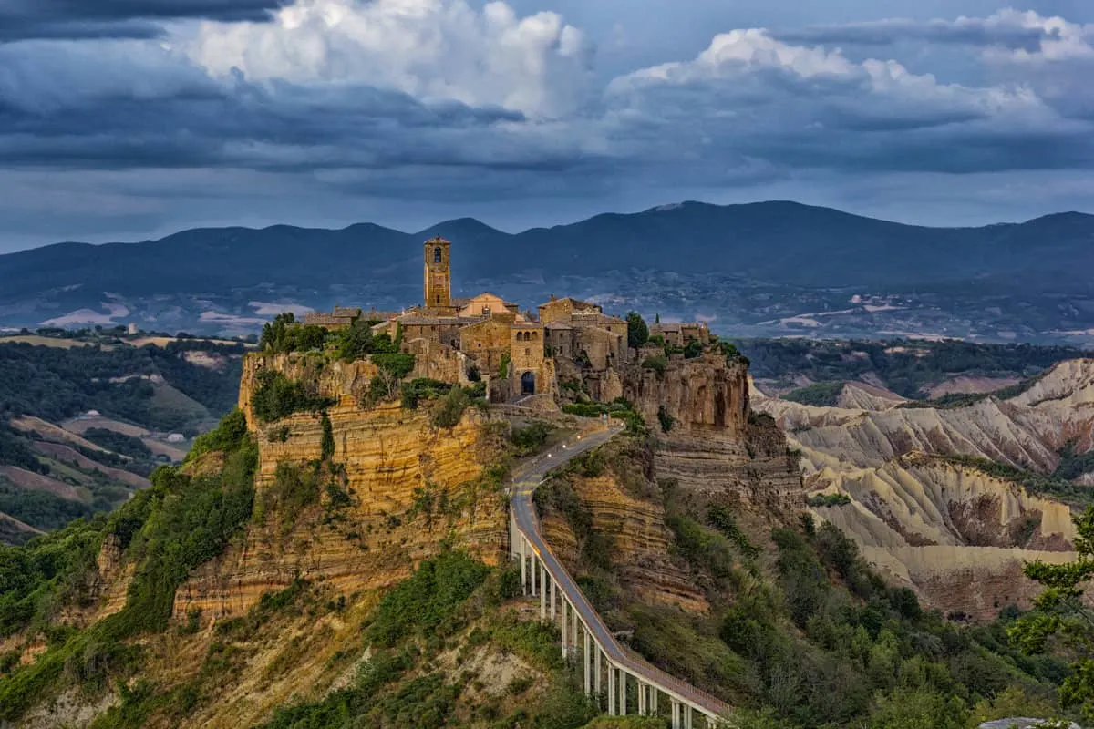 A Medieval hill town with a bridge leading to it and a spire at the top of the town. It is a dark and stormy sky.