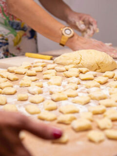 A wooden tray of hand rolled gnocchi. A womans hands in the distance rolling dough.