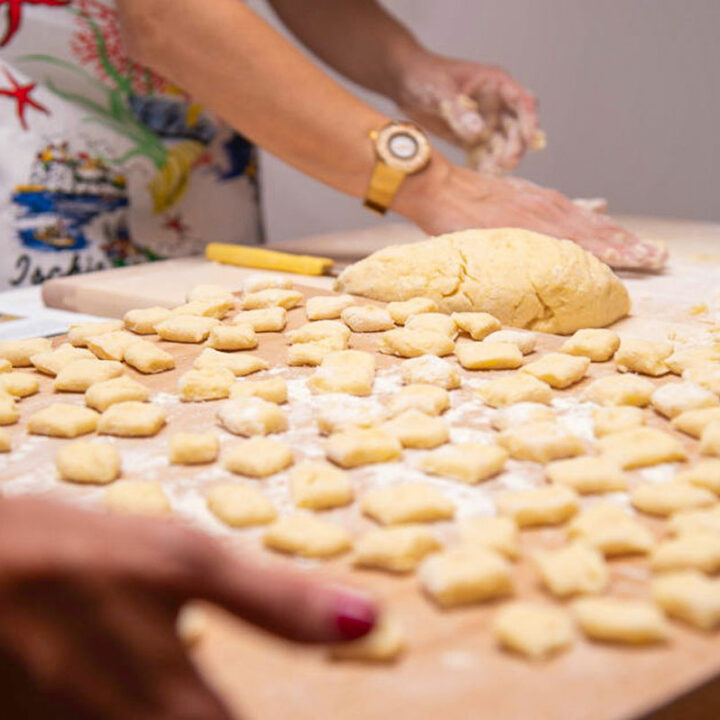 A wooden tray of hand rolled gnocchi. A womans hands in the distance rolling dough.