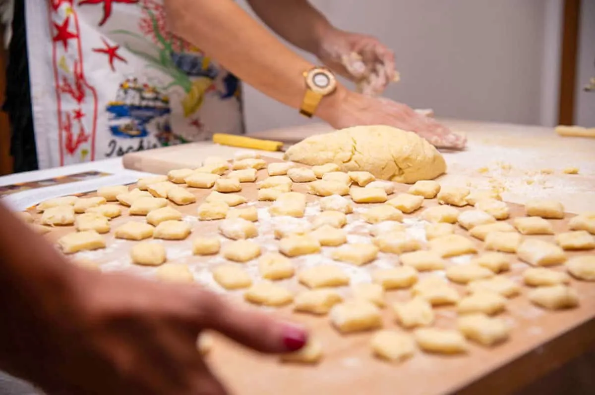 A wooden tray of hand rolled gnocchi. A womans hands in the distance rolling dough.