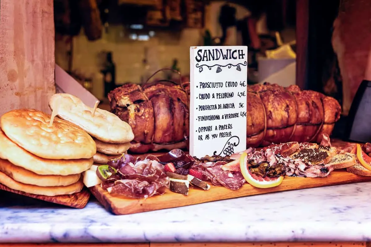 A snadwich shop with Italian bread and cured meats with a menu board. A large rolled pork sits behind the menu.