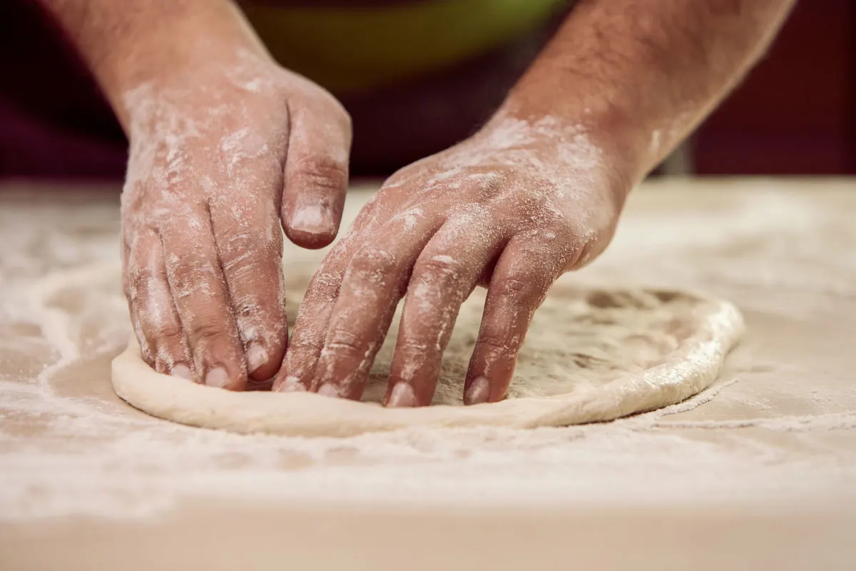 A mans floury hands gently press pizza dough into shape. 