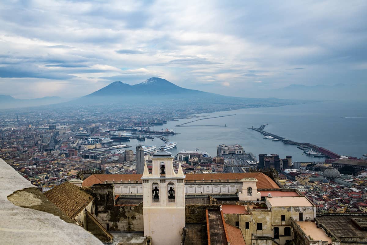 A view of the Bay of Naples with the city in the foreground and Mt Vesuvius in the distance on a cloudy day.