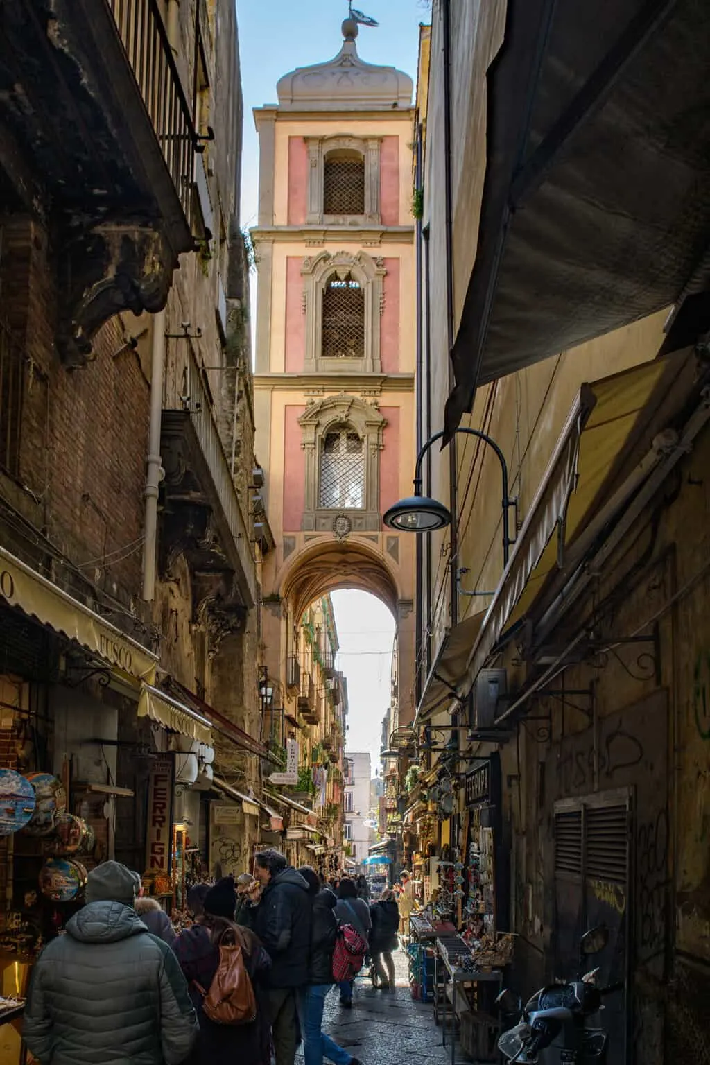 Tourists walk in a dark narrow street with souvenirs in Naples Italy, There is an arched steeple tower at the end of the street.