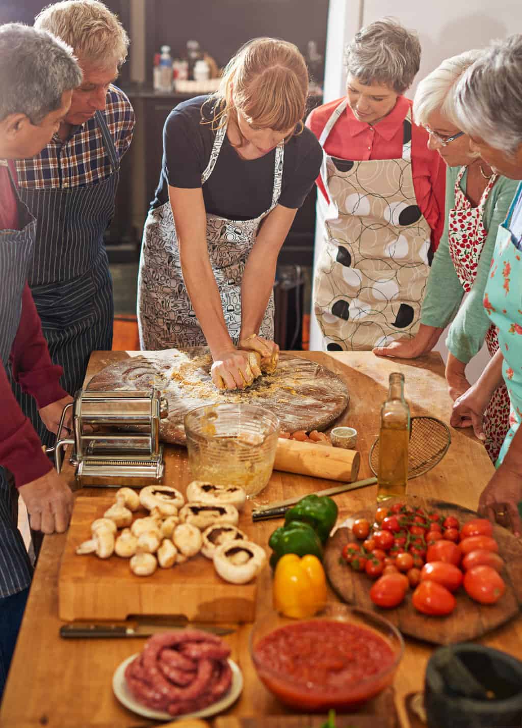 A group of senior peopl in aprons stand around a woman demonstrating pasta making. 