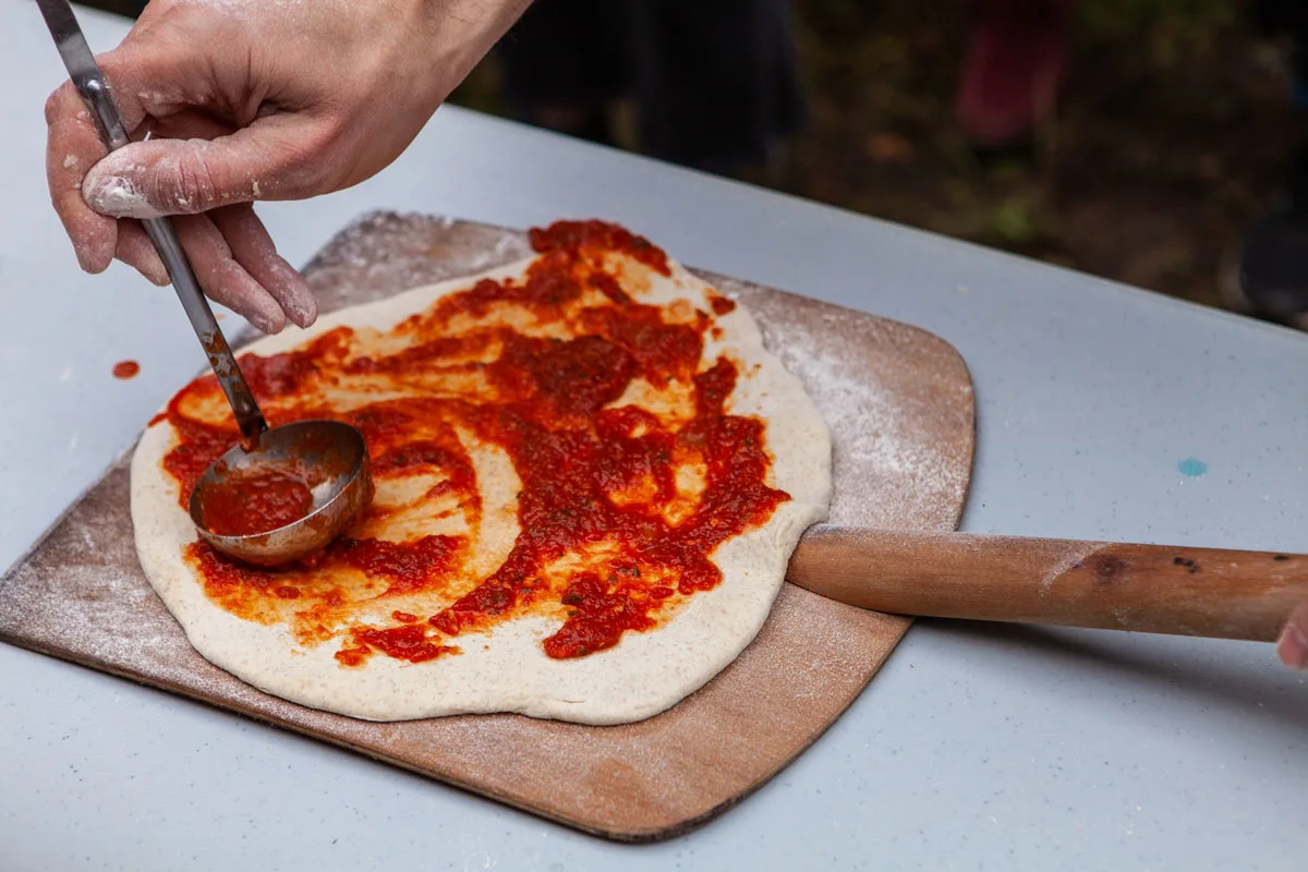 Man is spreading tomato paste on freshly kneaded pizza dough.