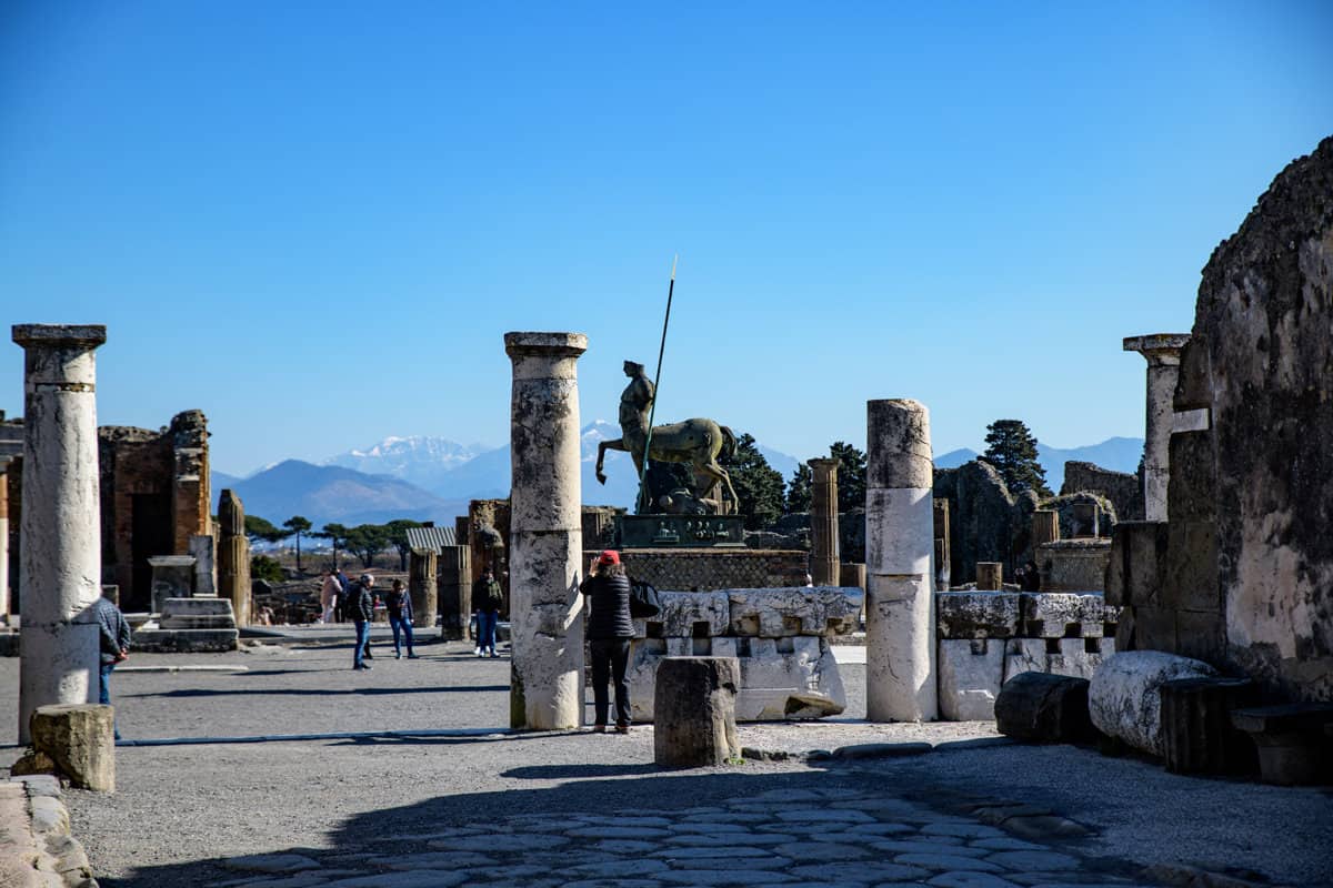 A tourists photographs the ancient ruins of Pompeii on a sunny day with snow capped mountains in the distance.