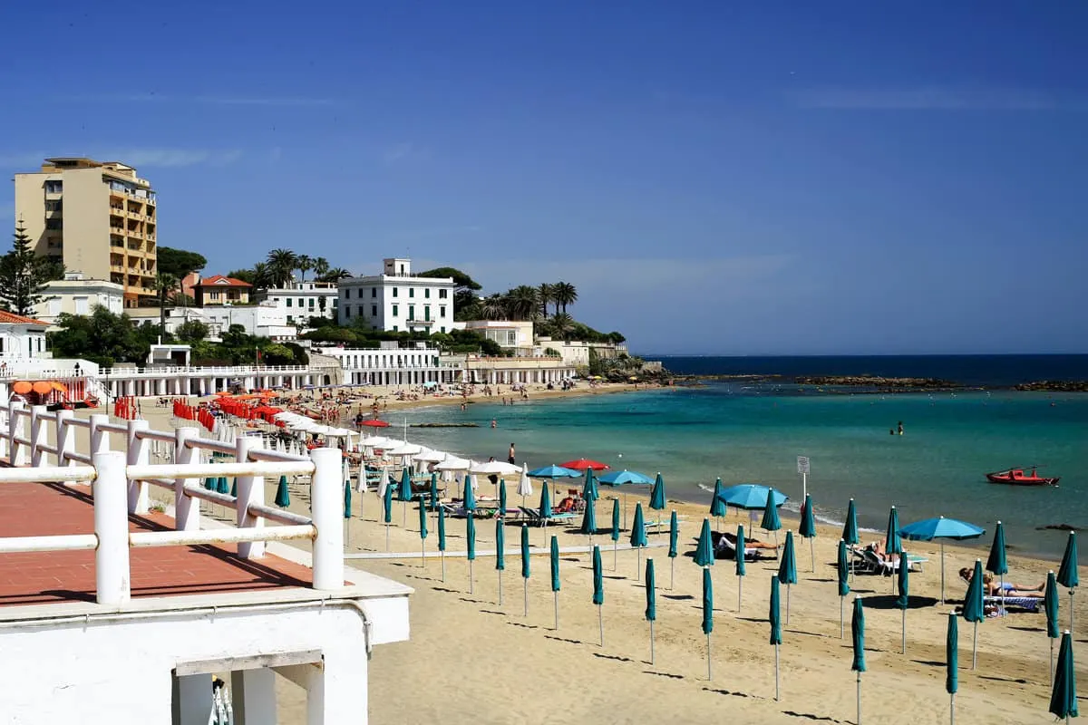 Beach umbrellas and sun lounges lined up on a white beach with clear water in an Italian coastal town.
