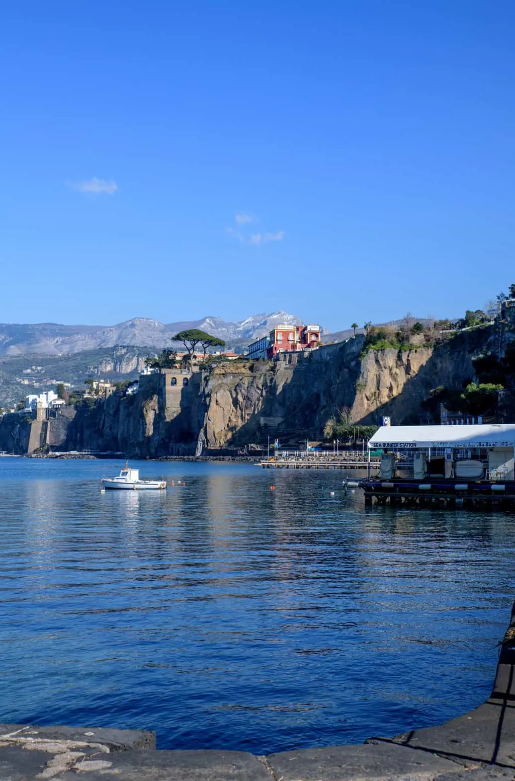 A boat on the harbour in front of cliffs. There is a large red colored house on top of the cliff.