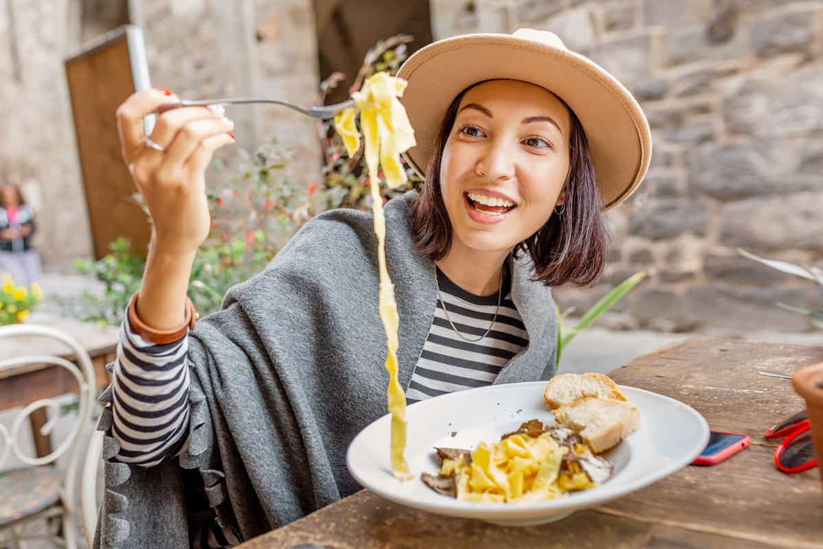 A happy asian woman wearing a hat holds up a string of pasta with her fork in a roman restaurant.