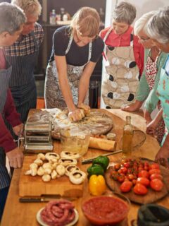 A group of senior peopl in aprons stand around a woman demonstrating pasta making.