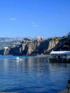 A boat on the harbour in front of cliffs. There is a large red colored house on top of the cliff.