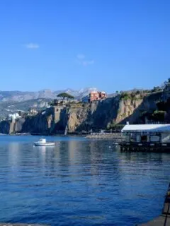 A boat on the harbour in front of cliffs. There is a large red colored house on top of the cliff.