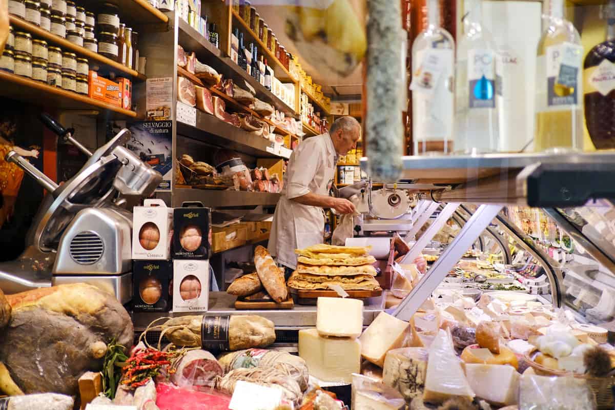 A man standing behind the counter of an Italian deli in Rome surrounded by cured meats and cheeses.
