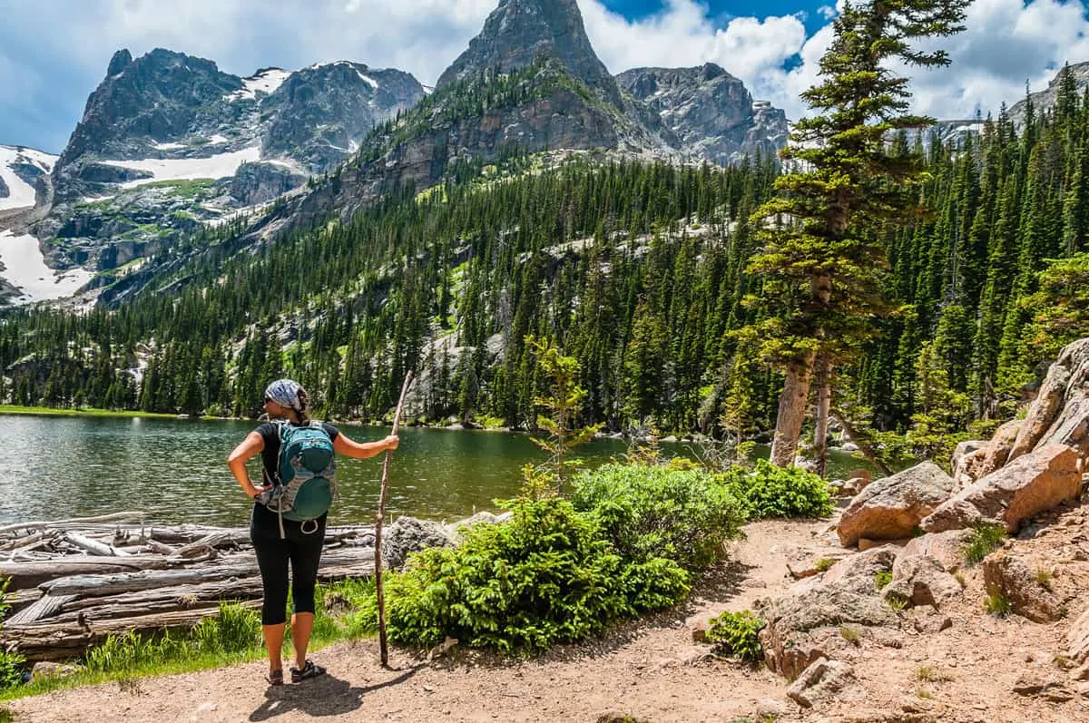 A female hiker stands in front of a lake on a sunny day with snow capped mountains on the other side of the lake.