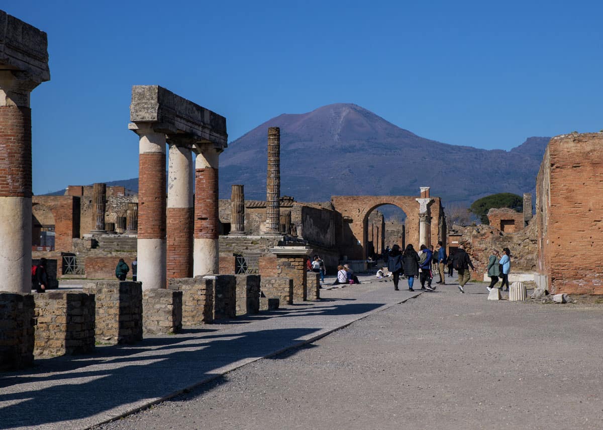 Tourists walking outisde on a sunny day around the ruins of Pompeii with Mount Vesuvius in the distance. 