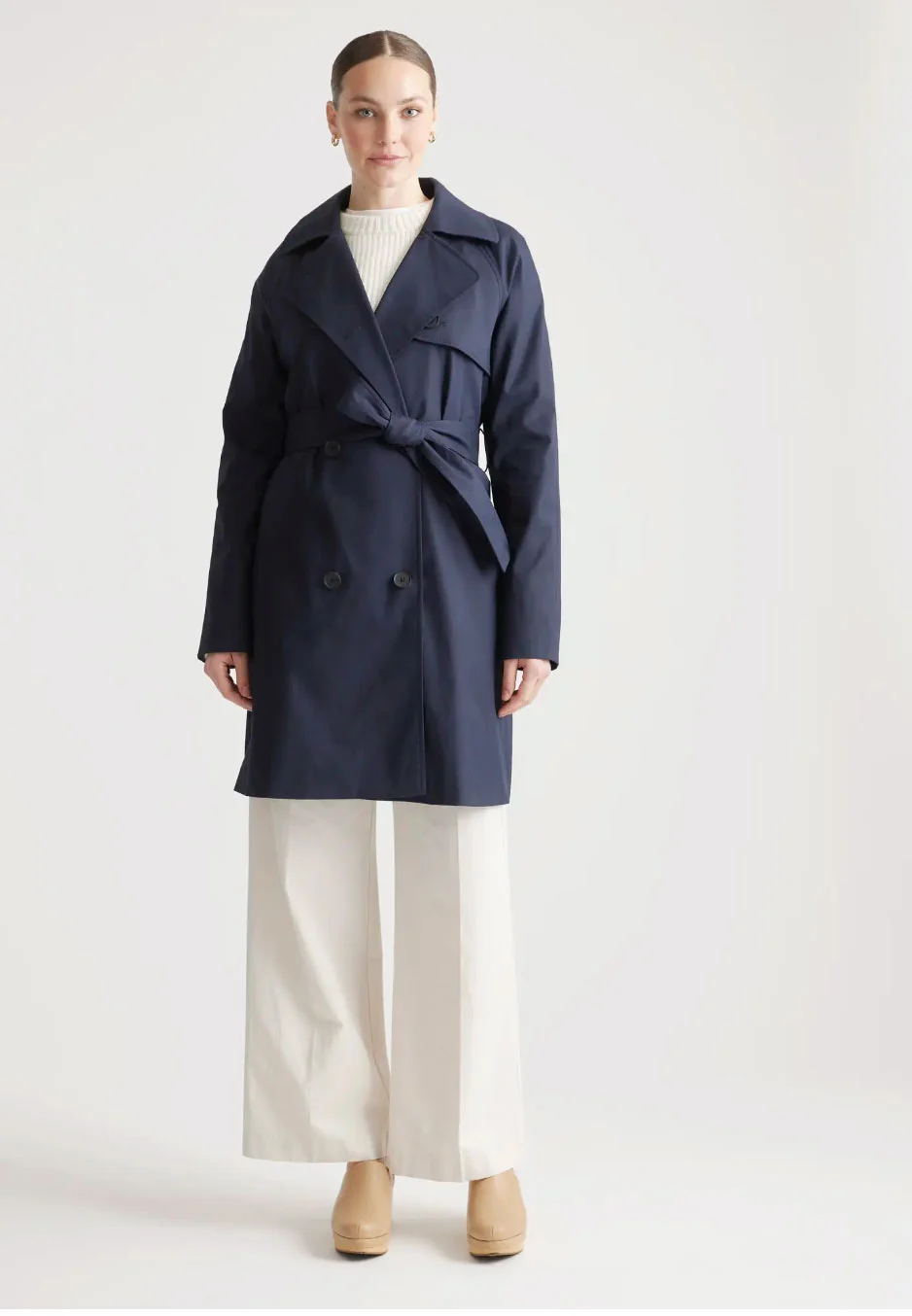 A blonde model wearing a navy travel trench with white pants in a white room. 