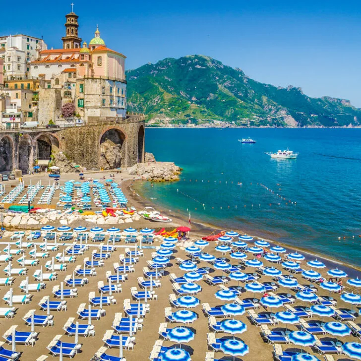 Blue and white beach umbrellas line the beach on the Amalfi Coast with pastel coloured buildings in the background.