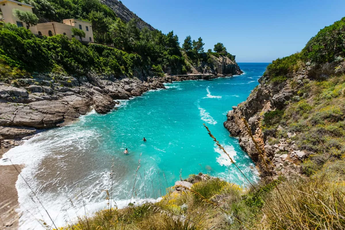 Two people swim in light blue clear water framed by green, rocky cliffs on a sunny day. 