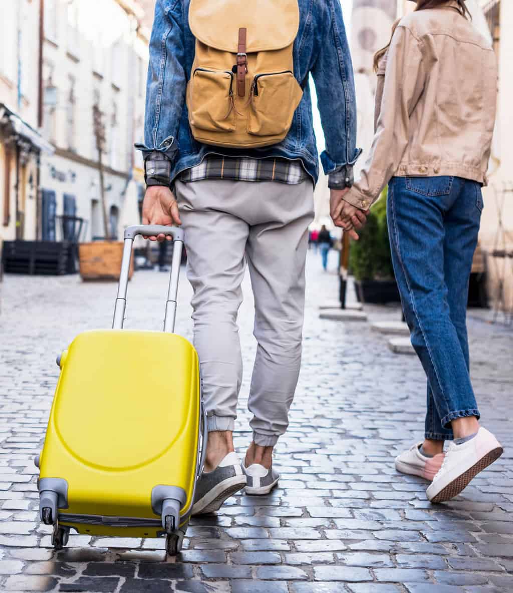 Cropped view of a couple walking down a cobbled street. He is pulling a yellow suitcase. They are holding hands. 