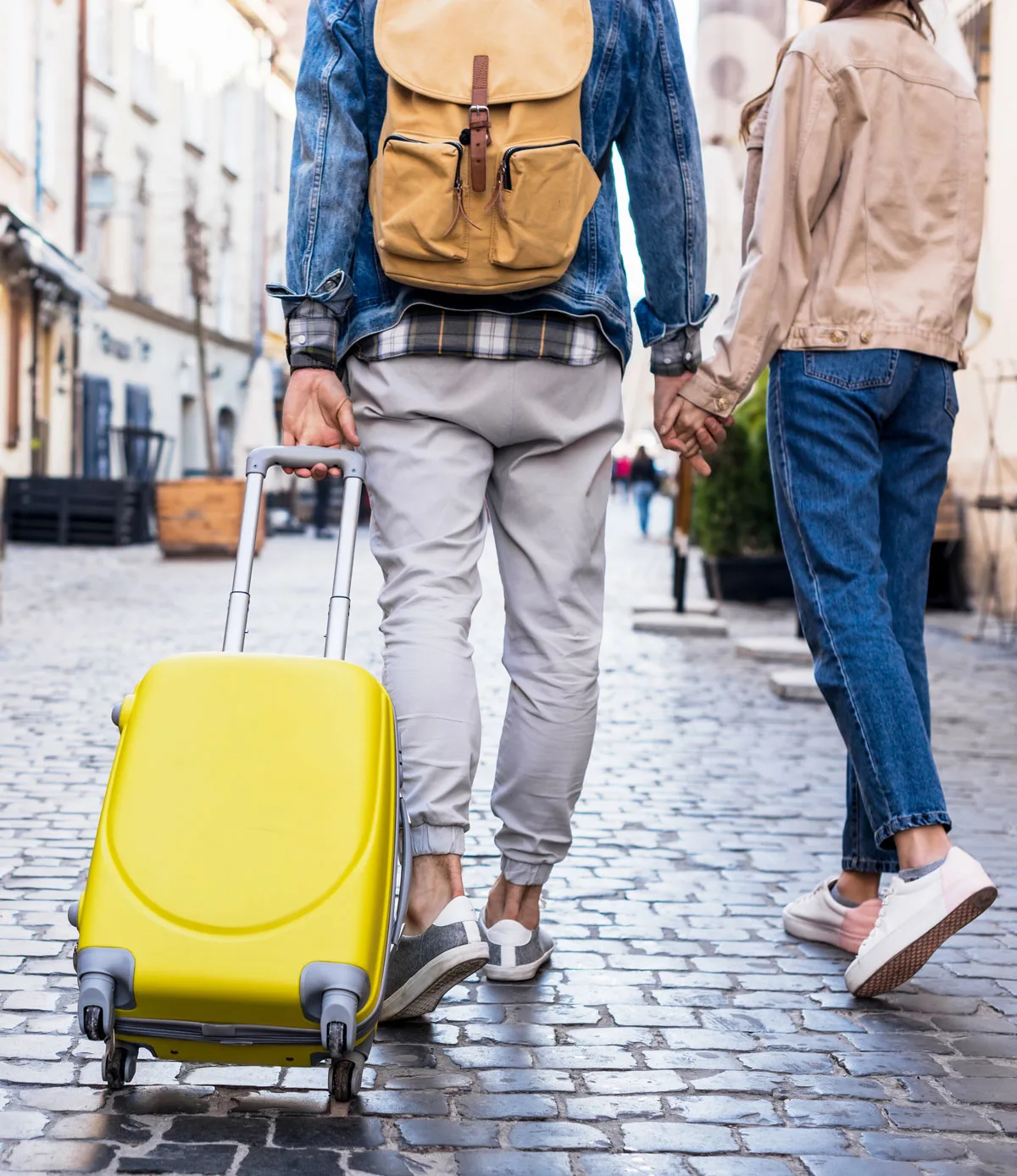 Cropped view of a couple walking down a cobbled street. He is pulling a yellow suitcase. They are holding hands.
