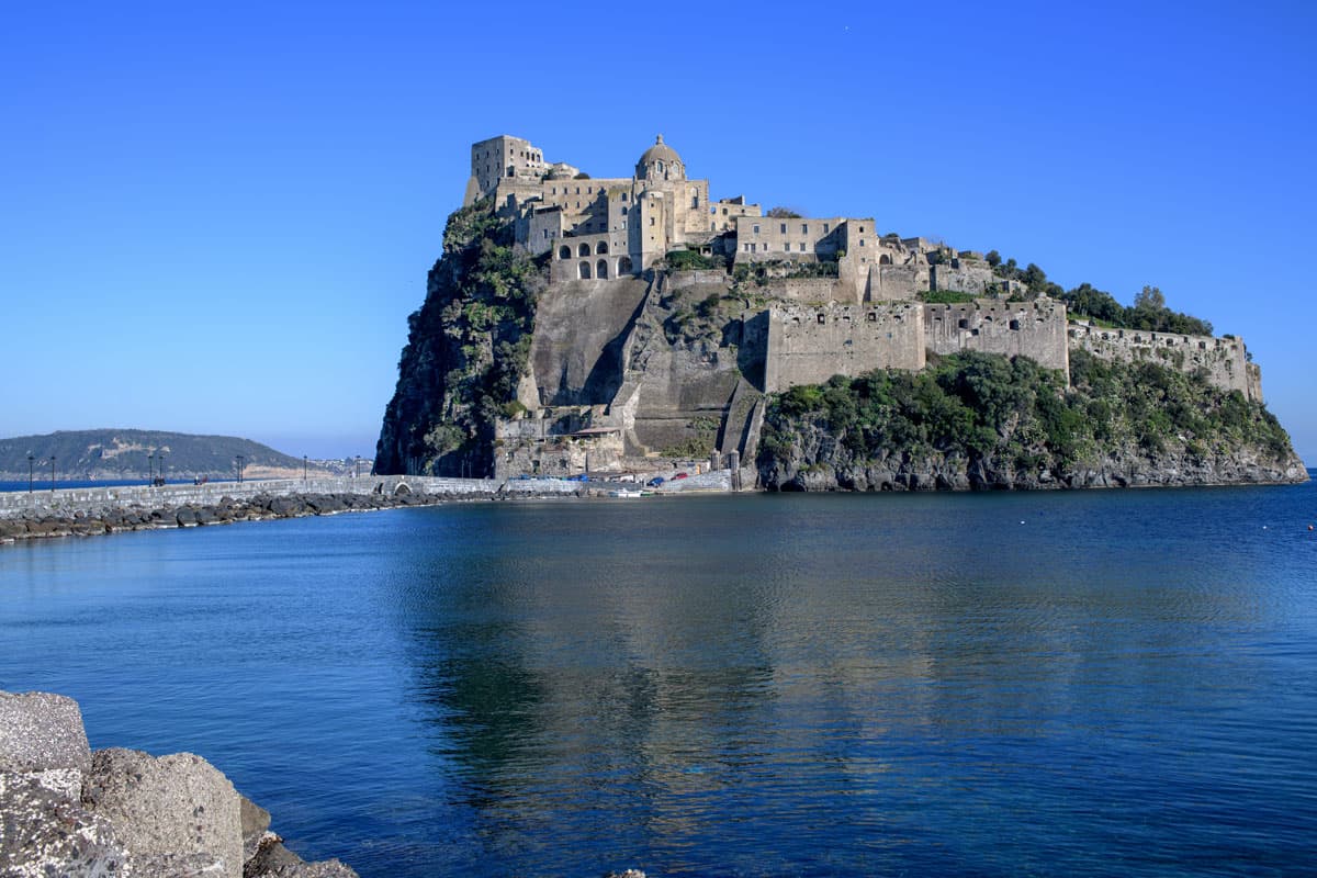An ancient castle sits on a rock island join by a bridge over blue water. 