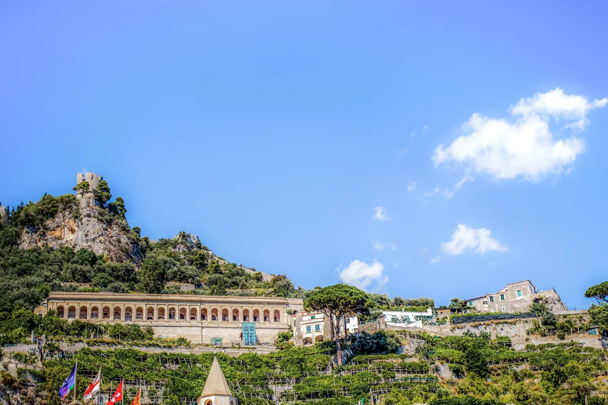 A watch tower rises above buildings in the green hills of the Amalfi Coast on a sunny day. 