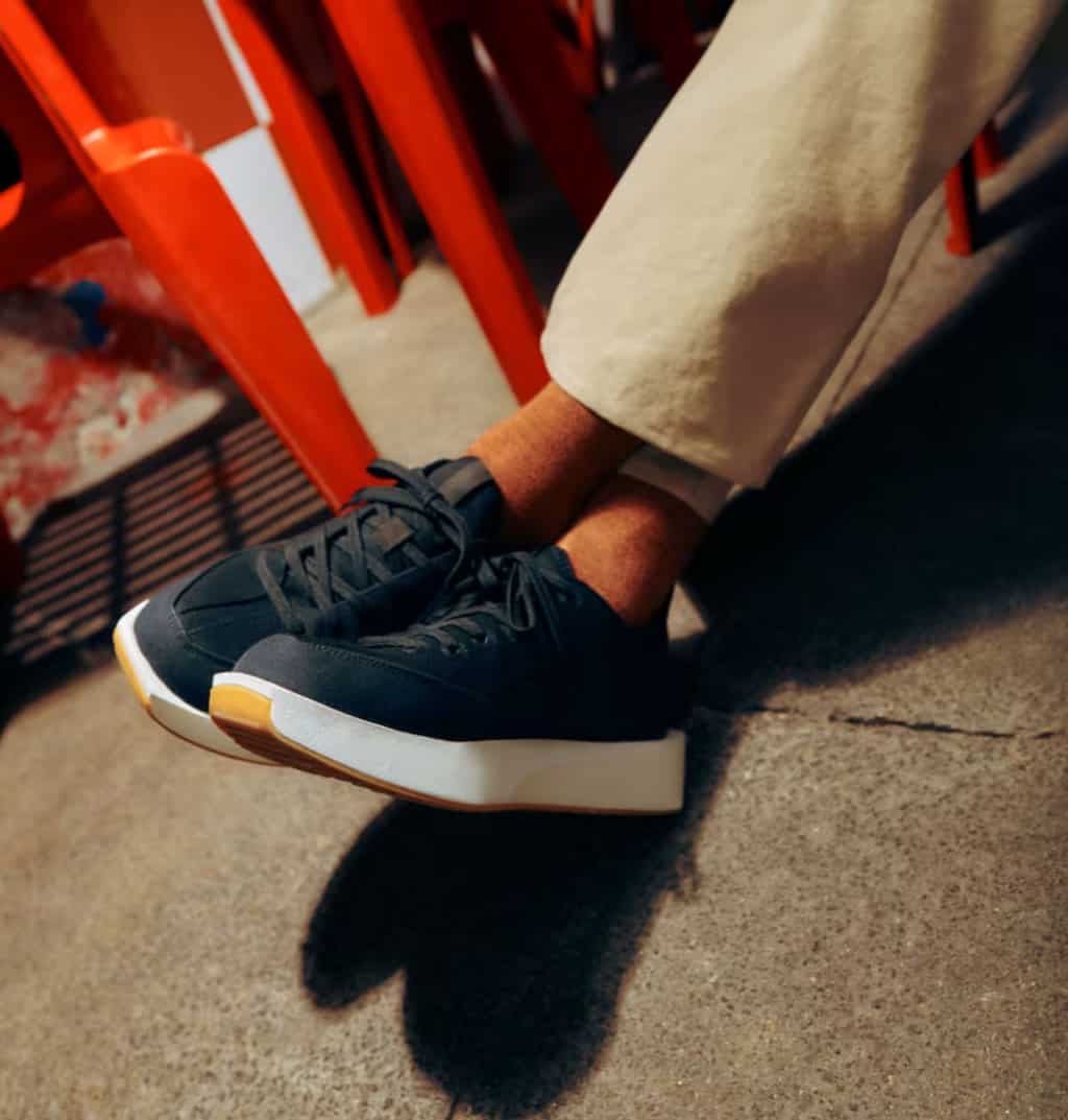 A mans feet crossed at the ankle wearing a pair of navy sneakers with tan pants. 