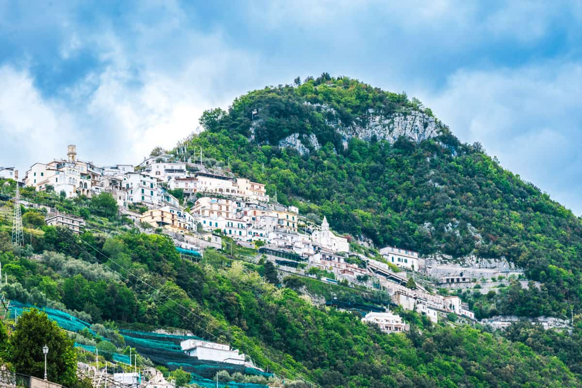 A white village cascades down a lush green mountain with a dramatic cloudy sky. 