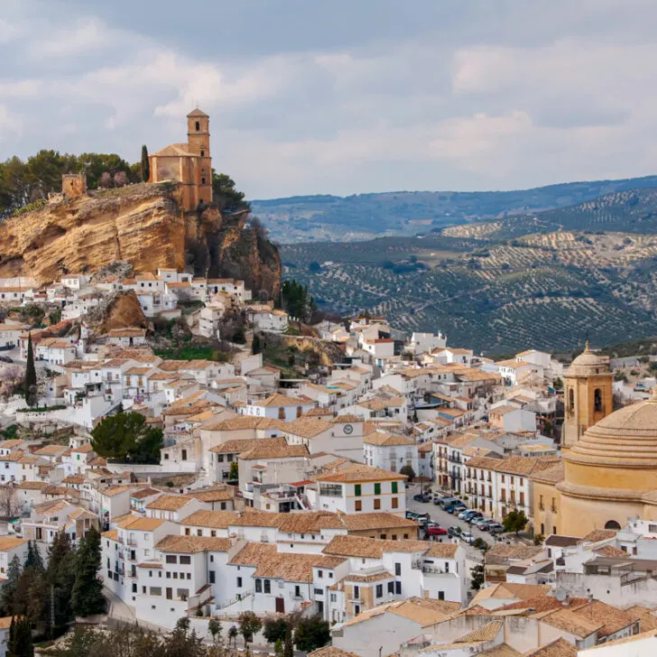 A small village of white houses spills down the hill crowned with a castle at the top of the hill in the countryside of southern Spain.