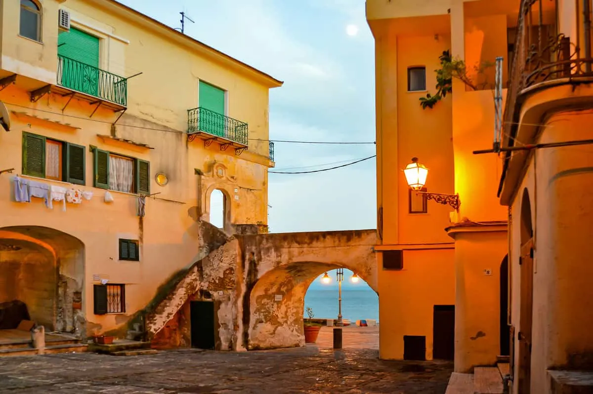 An old coastal fishing town glows yellow in the dusk. the sea is visible through a stone arch.