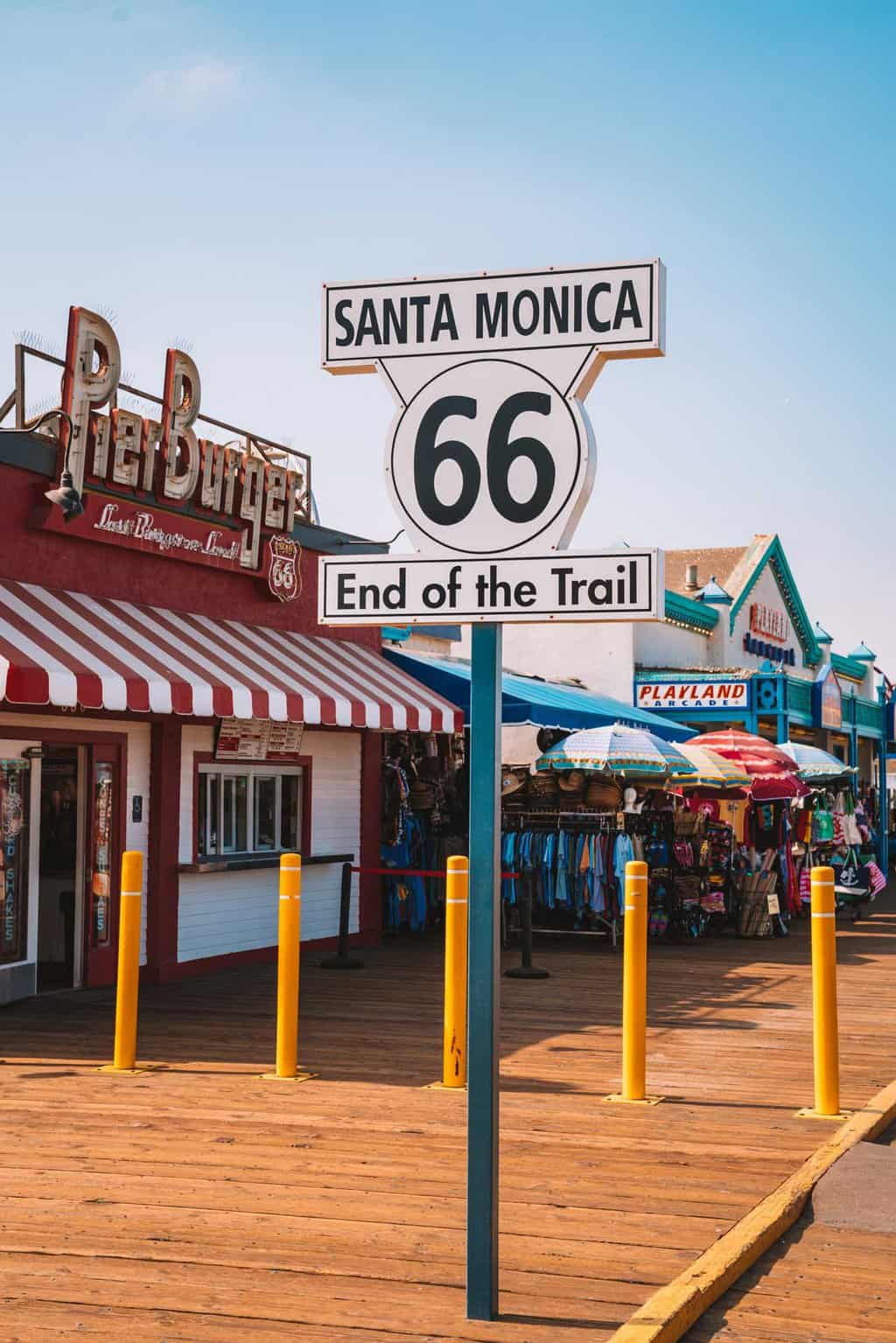 Santa Monica Route 66 End of the Trail sign on a boardwalk in front of colourful shops and restaurants. 