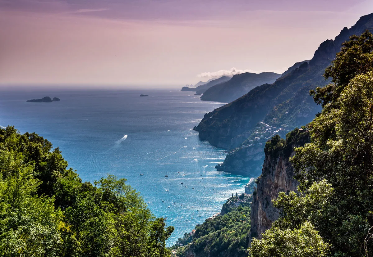 Looking down thrugh steep mountains over the sea where boats move about leaving a white trail. Sunset casts a purple glow over the Amalfi Coast.