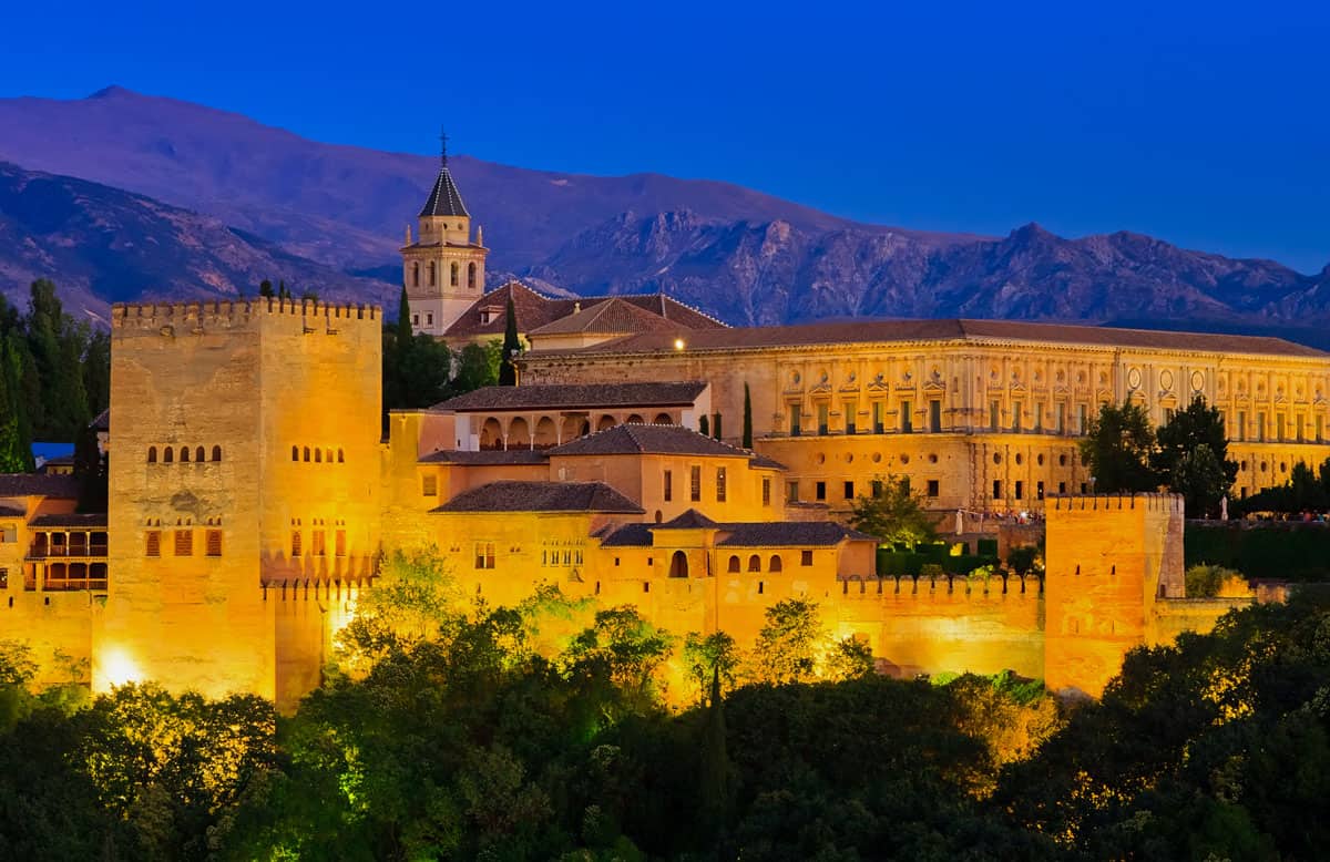 The Alhambra palace in Granada lit up at night. It glows golden against the dark mountain backdrop. 