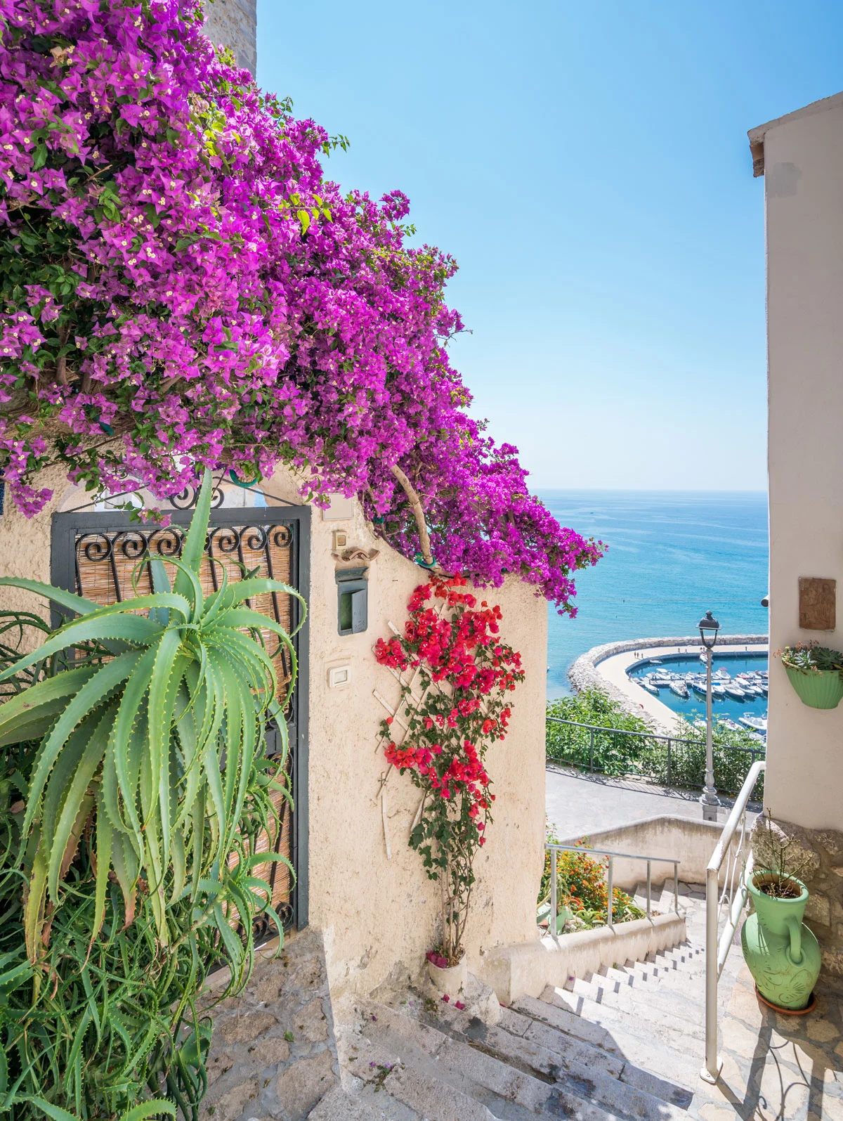 A stairway flanked with vibrant flowers and plants overlooking blue water and a boat marina in the Mediterranean. 