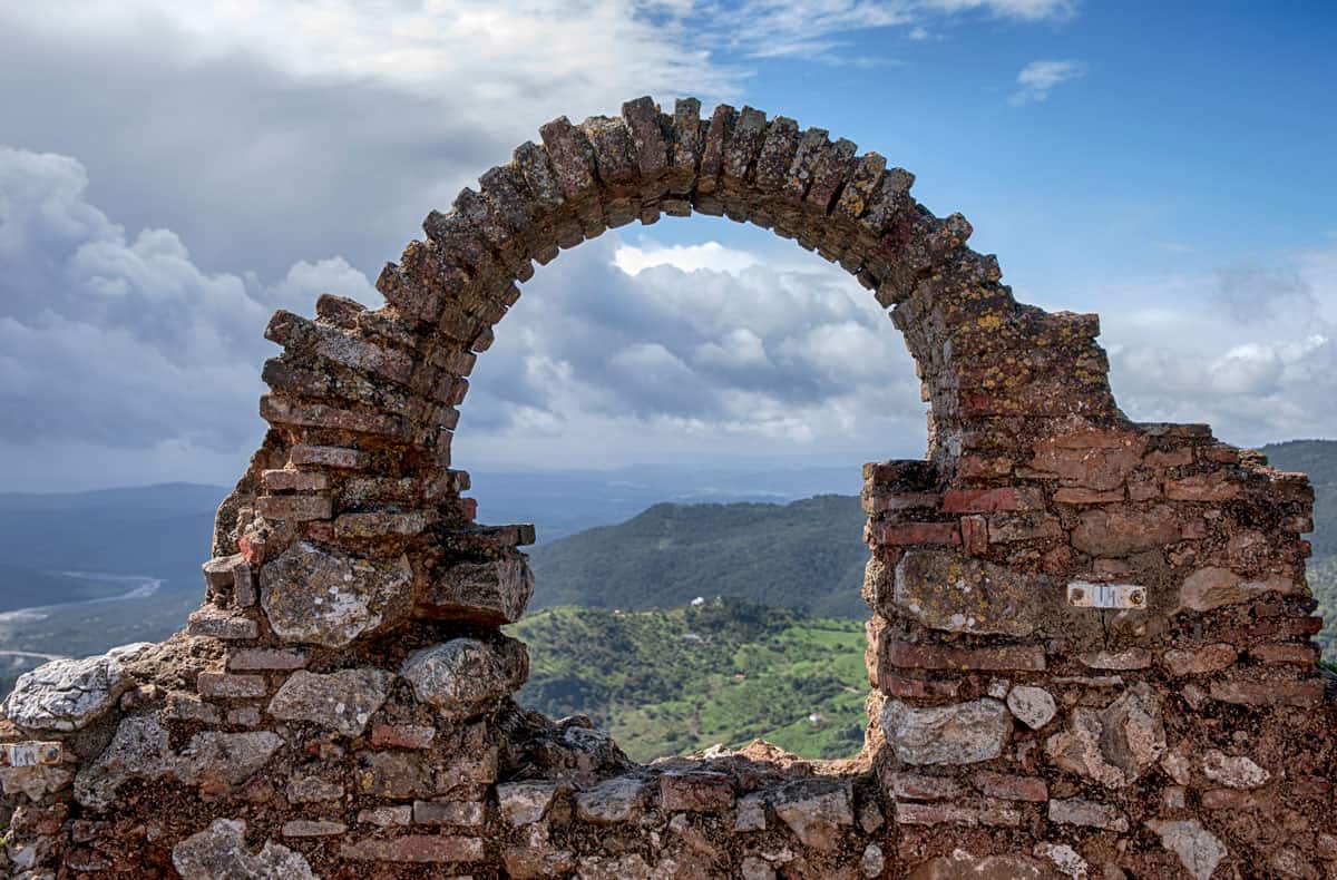 Looking through an archway of a castle ruin across the Spanish countryside with a dramatic sky. 